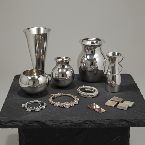 Nature morte présentant des objets d’art en argent, des bijoux, des récipients sculpturaux et des échantillons d’alliages métalliques sur une table sombre.